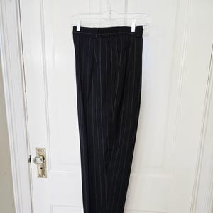 Puede incluir: Pantalones negros con finas rayas blancas, colgados de una percha transparente. Los pantalones tienen un diseño clásico con rayas verticales. La tela oscura cuelga de una puerta blanca.
