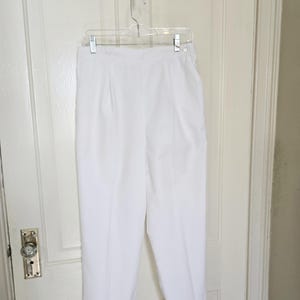 May include: White pants hanging on a clear hanger. The pants are made of a lightweight fabric and have a pleated front. The pants are a solid white color and appear to be a casual style.