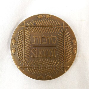 Puede incluir: Una medalla de bronce con una inscripci&oacute;n en hebreo que dice "Sukkot" y la palabra inglesa "Succot" en un marco decorativo.