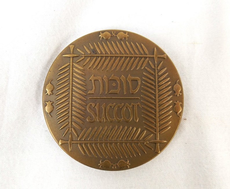 Puede incluir: Una medalla de bronce con una inscripci&oacute;n en hebreo que dice "Sukkot" y la palabra inglesa "Succot" en un marco decorativo.