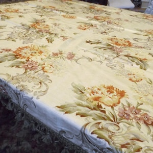 Puede incluir: Un mantel de color crema con un patrón floral con flores naranjas, amarillas y rosas. El mantel tiene un borde con flecos.