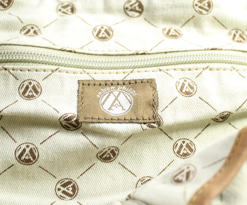May include: Close-up of a bag's interior. The light beige lining features a repeating pattern of brown circles and diagonal lines. A brown label with "andrew phillips" is visible.