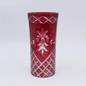 Puede incluir: Un vaso alto de color rojo rubí con intrincados patrones florales y geométricos grabados. El vaso presenta un patrón en forma de diamante en la base y un diseño floral en el centro. El borde tiene un diseño entrecruzado.