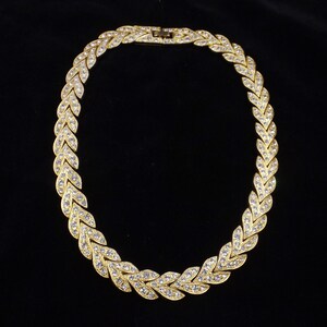 Puede incluir: Un collar de tono dorado con un diseño trenzado adornado con diamantes de imitación transparentes.