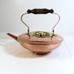 May include: A copper teapot with a brass lid and a wooden handle. The teapot has a wide, shallow body and a spout on the side.