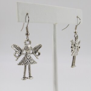 K&ouml;nnte beinhalten: Silberne Ohrh&auml;nger mit einem kleinen, detaillierten Anh&auml;nger einer M&auml;dchenfigur mit Fl&uuml;geln und Herzen auf ihrem Kleid.