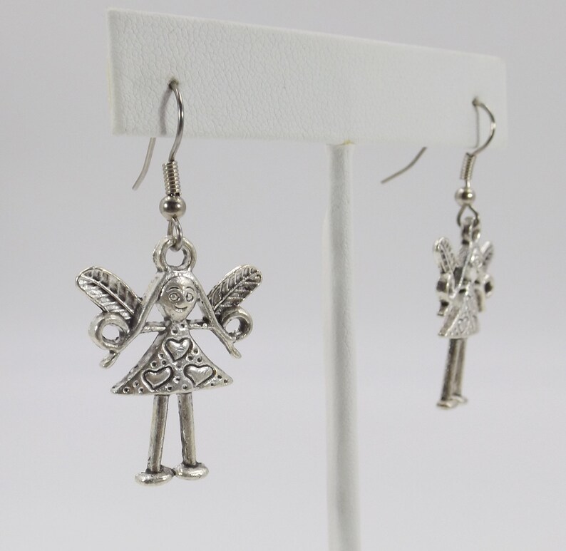 K&ouml;nnte beinhalten: Silberne Ohrh&auml;nger mit einem kleinen, detaillierten Anh&auml;nger einer M&auml;dchenfigur mit Fl&uuml;geln und Herzen auf ihrem Kleid.