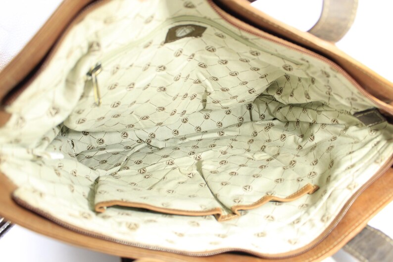 May include: An open tan leather tote bag. The interior is lined with light green fabric patterned with a repeating emblem. A zipped pocket and other pockets are visible. The bag has a rectangular shape.