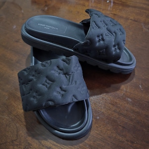May include: Black slide sandals with a quilted upper featuring a repeating logo pattern. The sandals have a thick sole and the brand name "LOUIS VUITTON PARIS" is visible on the footbed.
