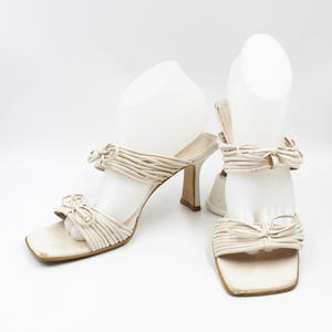 May include: A pair of white heeled sandals with a woven strap design and bow accents.