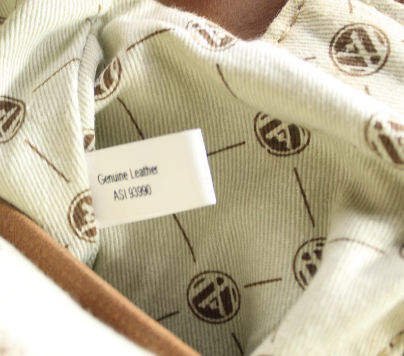 May include: Close-up of the interior of a brown leather bag. The lining is a light beige fabric with a repeating brown logo pattern. A white tag reads "Genuine Leather" and "ASI 93990".