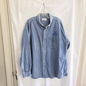 May include: Light blue denim button-down shirt with long sleeves. Features a chest pocket and the embroidered text "Allstate" in blue. The shirt is hanging on a hanger against a white backdrop.