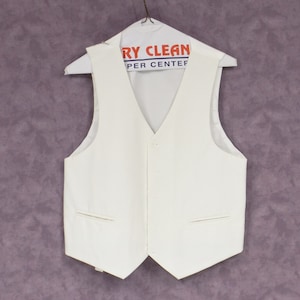 Puede incluir: Un chaleco blanco con cuello en V y un cierre de botón único. El chaleco tiene dos bolsillos en la parte delantera y está colgado en una percha con una etiqueta de limpieza en seco que dice "Dry Clean Per Center".