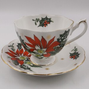 May include: A white teacup and saucer set featuring a Christmas poinsettia design. The cup and saucer are decorated with red poinsettias, green holly leaves, and gold stars.