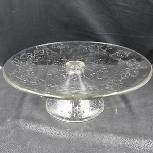Puede incluir: Un soporte para pasteles de vidrio transparente con una parte superior redonda y una base de pedestal. La superficie superior presenta un patrón floral. El soporte está diseñado para exhibir pasteles y otros postres. El soporte es de vidrio transparente.