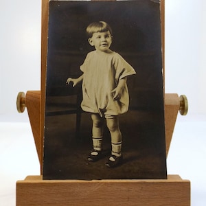 Puede incluir: Fotografía antigua en blanco y negro de un niño pequeño. El niño lleva un pelele de color claro, calcetines y zapatos Mary Jane. La fotografía se muestra en un caballete de madera.