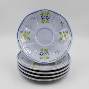 May include: A stack of six light blue ceramic saucers with a scalloped edge and a blue rim. Each saucer features a floral design with green, yellow, and purple accents. The saucers are stacked on top of each other.