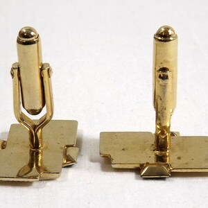 May include: A pair of gold-toned cufflinks with a geometric design. The cufflinks have a square base and a vertical post with a rounded top.