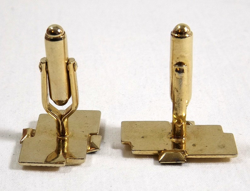 May include: A pair of gold-toned cufflinks with a geometric design. The cufflinks have a square base and a vertical post with a rounded top.