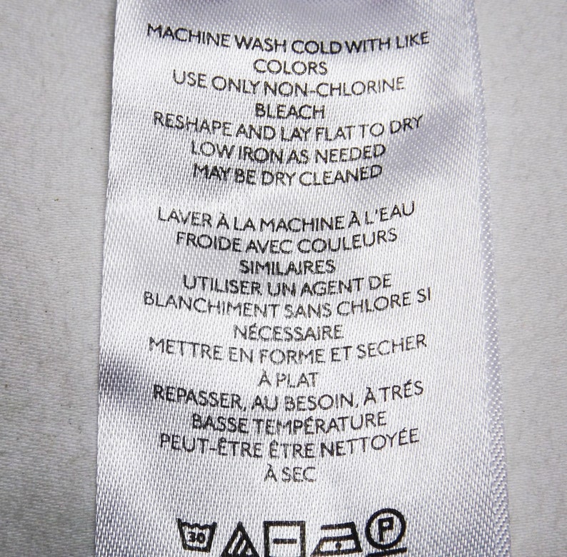 May include: White care label with instructions for washing, drying, and ironing a garment. The label includes both English and French instructions. The text reads "Machine wash cold with like colors. Use only non-chlorine bleach. Reshape and lay flat to dry. Low iron as needed. May be dry cleaned. Laver &agrave; la machine &agrave; l'eau froide avec couleurs similaires. Utiliser un agent de blanchiment sans chlore si n&eacute;cessaire. Mettre en forme et s&eacute;cher &agrave; plat. Repasser, au besoin, &agrave; tr&egrave;s basse temp&eacute;rature. Peut-&ecirc;tre &ecirc;tre nettoy&eacute;e &agrave; sec."