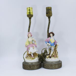 Op de afbeelding: Twee vintage porseleinen lampen met figuren in historische kleding. De lampen hebben messing stelen en sierlijke voetstukken. Eén lamp toont een vrouw in een roze en gele jurk, de andere een man in blauwe en roze kleding. Op een witte achtergrond.