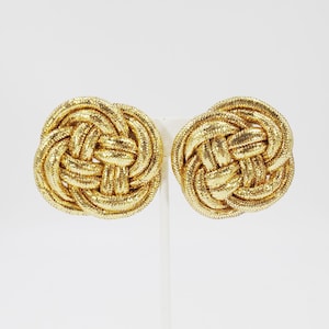 Peut inclure: Une paire de boucles d'oreilles à clip dorées en forme de nœud tressé. Les boucles d'oreilles sont rondes et présentent un motif tressé texturé. Les boucles d'oreilles sont présentées sur un fond blanc.