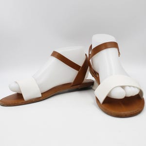 Könnte beinhalten: Ein Paar weiße und braune Ledersandalen mit Knöchelriemen. Die Sandalen haben eine flache Sohle und ein schlichtes Design.