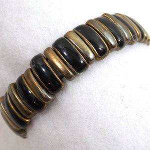 Puede incluir: Un brazalete negro y dorado hecho de cuentas ovaladas enlazadas. Las cuentas alternan negro y dorado.