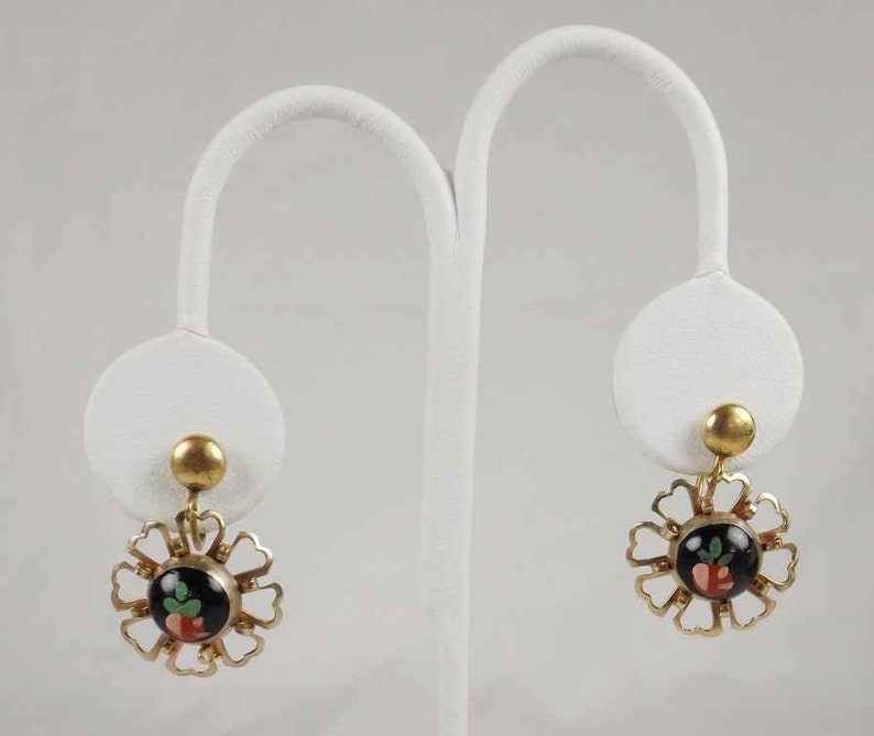 May include: A pair of gold tone earrings with a black and red floral design. The earrings are on a white display stand.