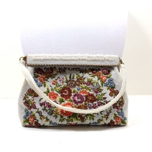Puede incluir: Un bolso blanco de cuentas con un diseño floral. El bolso tiene un cierre dorado y una correa blanca de cuentas.