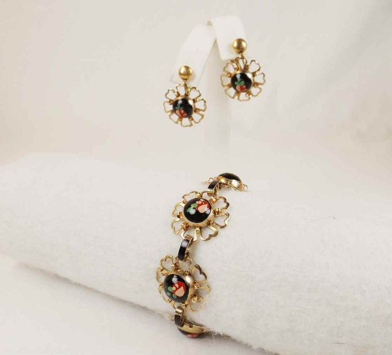 May include: A gold bracelet and earrings set with black enamel and floral accents. The bracelet has five round black enamel charms with floral designs in red, white, and green. The earrings are drop earrings with a similar round black enamel charm with floral accents.