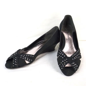 Puede incluir: Un par de zapatos de tacón de cuña negros de punta abierta con un lazo de pedrería. Los zapatos son de la marca "Renee".