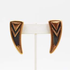 May include: A pair of gold-toned, geometric earrings. Each earring has a curved, elongated shape with a dark blue-green center and a series of layered, triangular designs. The earrings are displayed on a white earring stand.