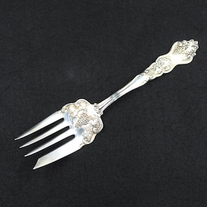 May include: A silver fork with an ornate handle featuring a grapevine design. The fork has four tines and a polished finish.