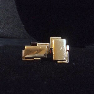May include: Two gold-colored cufflinks with a red border. The cufflinks are rectangular and have a textured surface.