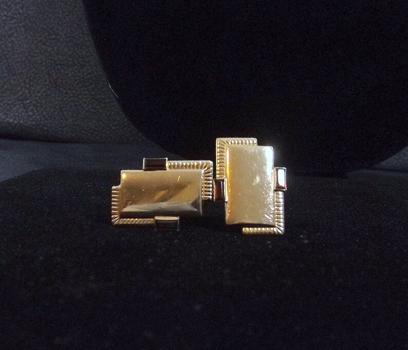 May include: Two gold-colored cufflinks with a red border. The cufflinks are rectangular and have a textured surface.