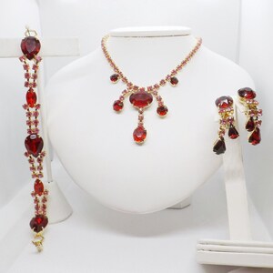May include: A set of red and pink rhinestone jewelry, including a necklace, bracelet, and earrings. The necklace features a teardrop-shaped pendant with a large red rhinestone.