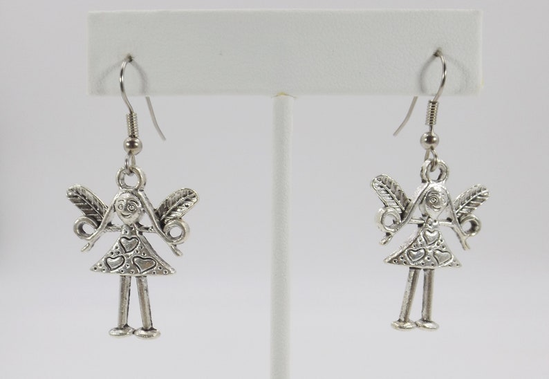K&ouml;nnte beinhalten: Silberne Ohrh&auml;nger mit einem kleinen, detaillierten Anh&auml;nger einer M&auml;dchenfigur mit Fl&uuml;geln und Herzen auf ihrem Kleid.