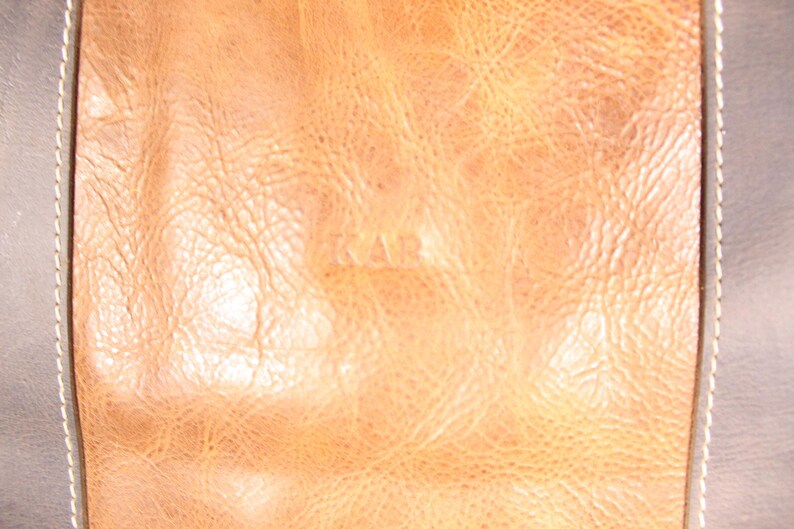 May include: Close-up of a section of leather. A central tan panel with a textured surface and the embossed letters "KAR" is bordered by darker, grey-brown leather with white stitching. The leather has a worn appearance.