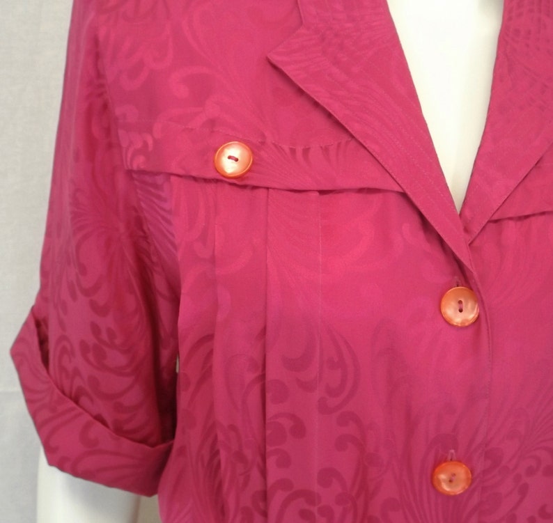 May include: A pink button-up shirt with a floral pattern and orange buttons. The shirt has a collar and short sleeves.
