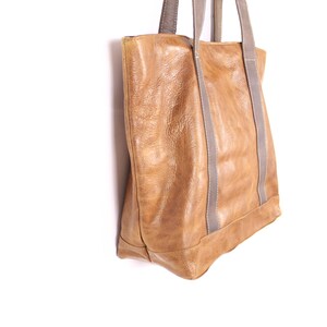 May include: A tan leather tote bag with grey straps. The bag has a rectangular shape with a reinforced base and visible stitching. The leather has a textured appearance.
