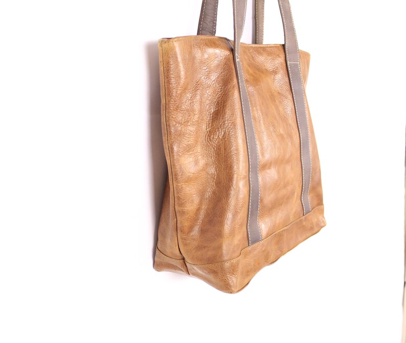May include: A tan leather tote bag with grey straps. The bag has a rectangular shape with a reinforced base and visible stitching. The leather has a textured appearance.