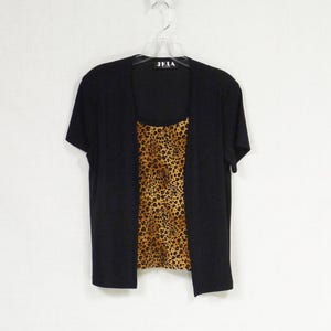 Puede incluir: Top negro de manga corta con un top de estampado de leopardo debajo. El top tiene el texto "JKLA" en la parte delantera.