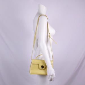 May include: A pale yellow handbag with a top handle and a long, adjustable shoulder strap. The bag has a flap closure with a gold-colored circular clasp. The bag is displayed on a white mannequin.