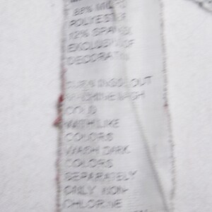May include: A white care label with black text detailing the fabric composition, 88% Microfiber Polyester, 12% Spandex, excluding decoration. The label also includes instructions for washing and drying the garment: cold wash like colors, wash dark colors separately, only non-chlorine bleach when needed, tumble dry low.