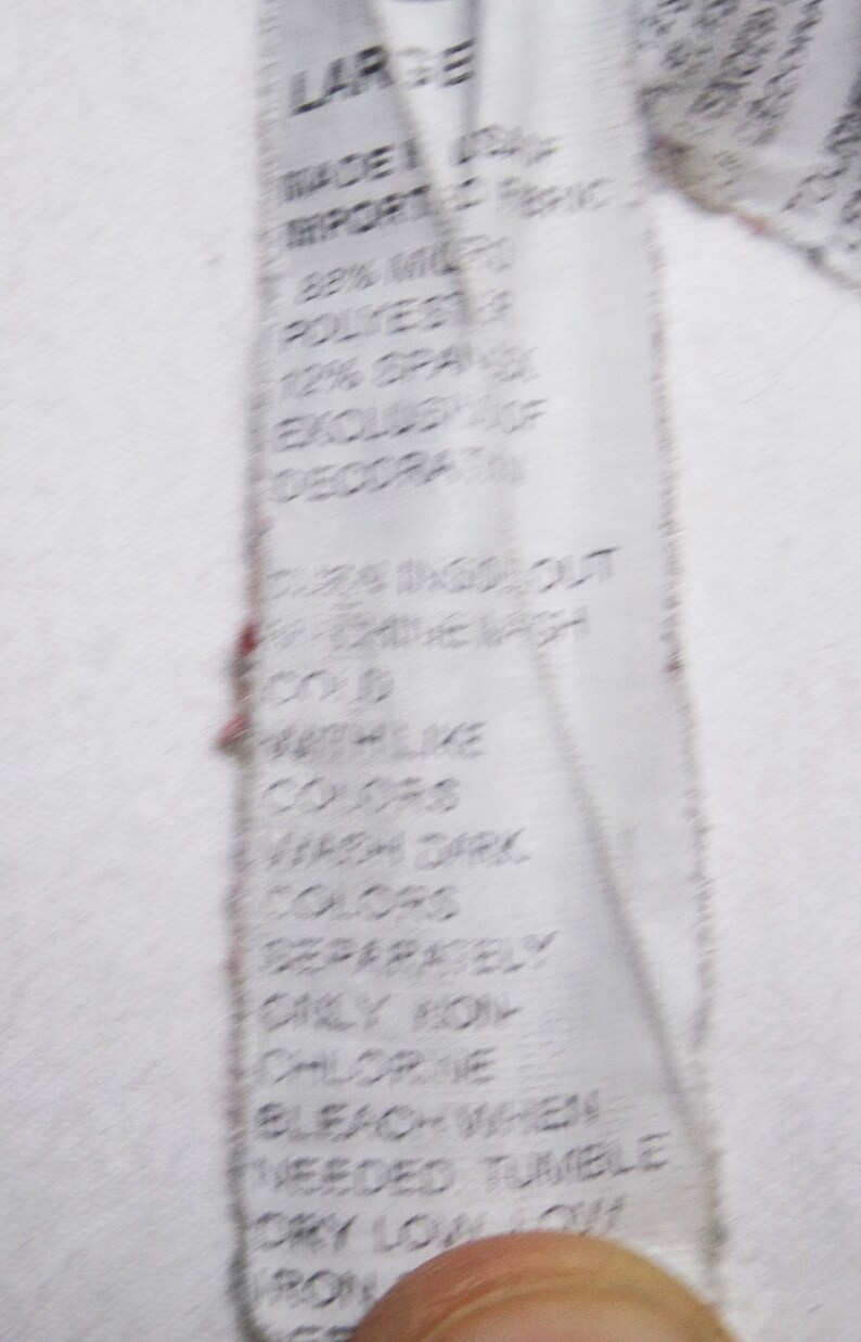 May include: A white care label with black text detailing the fabric composition, 88% Microfiber Polyester, 12% Spandex, excluding decoration. The label also includes instructions for washing and drying the garment: cold wash like colors, wash dark colors separately, only non-chlorine bleach when needed, tumble dry low.