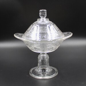 May include: A clear glass pedestal dish with a lid. The dish has a decorative pattern and is on a pedestal base.