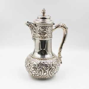 Könnte beinhalten: Eine silberne Kanne mit einem verzierten Blumenmuster. Die Kanne hat einen runden Körper und einen hohen, schlanken Hals. Der Griff ist gebogen und hat ein dekoratives Design. Die Kanne ist aus poliertem Silber gefertigt und hat eine glänzende Oberfläche.