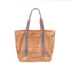 May include: A large, rectangular tote bag in a warm, tan colour. The bag features two long, grey handles with a contrasting stitch detail. The bag appears to be made of leather or a similar material, with a slightly distressed finish.