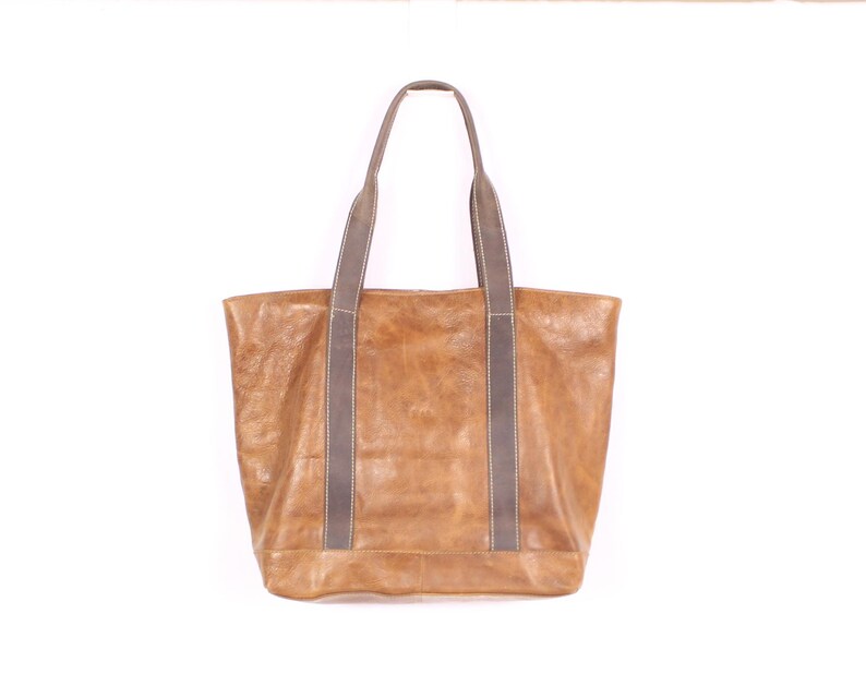 May include: A large, rectangular tote bag in a warm, tan colour. The bag features two long, grey handles with a contrasting stitch detail. The bag appears to be made of leather or a similar material, with a slightly distressed finish.